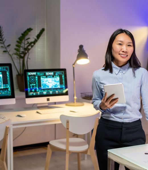 Portrait of smiling attractive Asian car designer in stripped shirt holding digital tablet in modern office with computers
